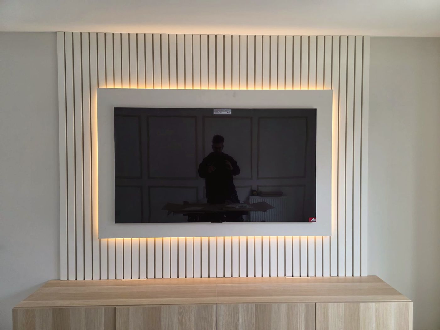 A white slatted media wall with warm LED backlighting around a wall-mounted TV and an oak floating unit